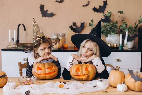 Mother and daughter wearing witch hats leaning on carved pumpkins on a table decorated for Halloween party