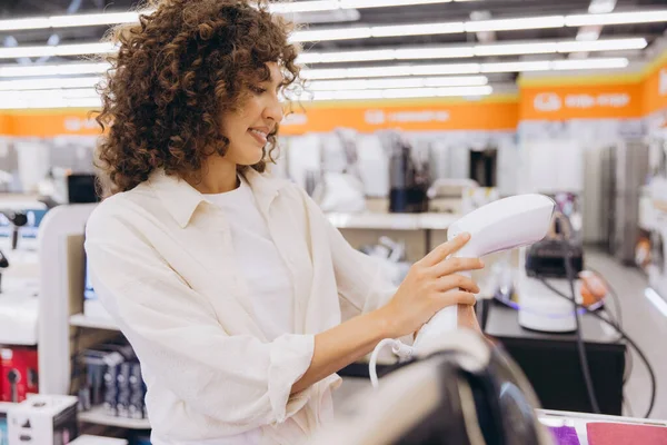 Smiling woman with curly hair selecting a new handheld garment steamer while browsing the shelves of an appliance store, enjoying the shopping experience