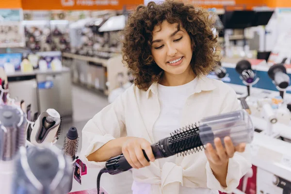 Smiling woman with curly hair selecting a new hair dryer while shopping in an electronics store filled with various appliances