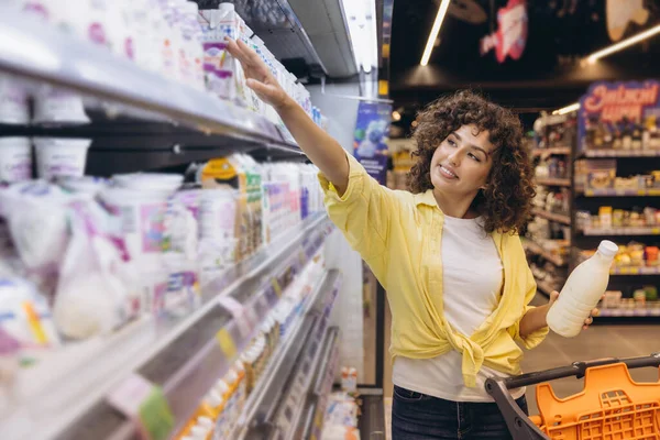 Customer selecting yogurt inside a grocery store refrigerator, holding a milk bottle and pushing a shopping cart