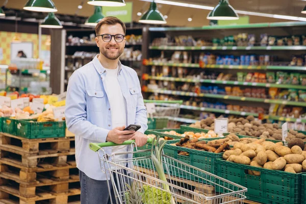 Man using smartphone and pushing shopping cart with fresh vegetables inside a supermarket
