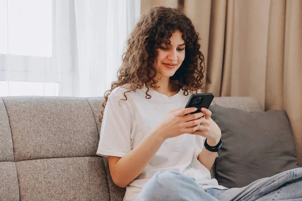Young woman using mobile phone at home, relaxing on comfortable couch, browsing internet, chatting online, shopping or watching video