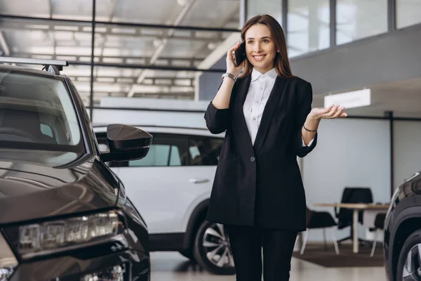 Smiling saleswoman in a suit engaging in a phone conversation while presenting a new car to an interested customer in the dealership showroom