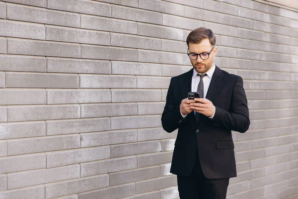 Businessman wearing suit and tie using smartphone standing near gray brick wall
