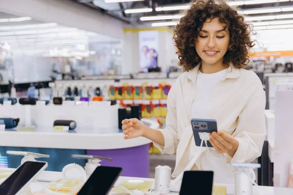 Smiling customer holding smartphone, comparing and choosing mobile phone in consumer electronics store