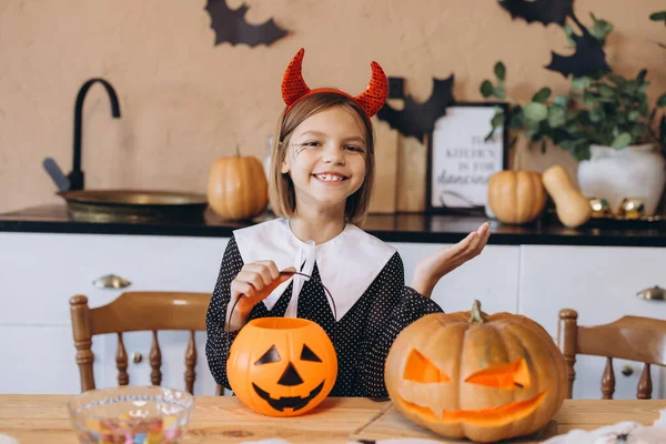 Smiling girl wearing devil costume showing halloween candy basket and carved pumpkin sitting at kitchen table