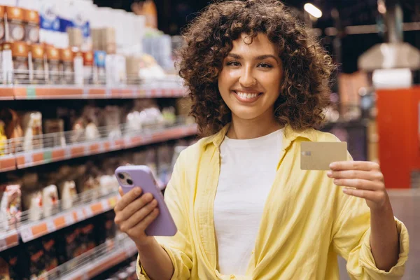 Young woman holding a smartphone and a credit card, smiling in a supermarket, ready for a convenient and modern shopping experience