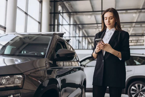 Saleswoman using a mobile phone while standing next to a car in a dealership showroom, engaging with customers and discussing options