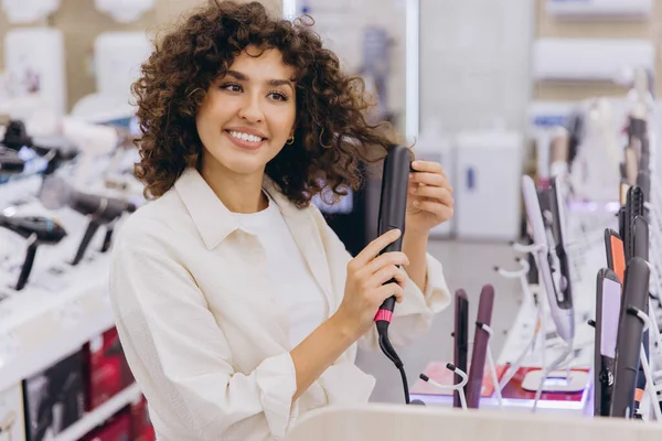 Smiling curly haired woman testing new hair straightener in an electronics store, trying different hair styling tools