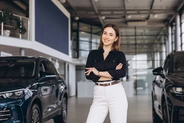 Saleswoman smiling with arms crossed while standing in a car dealership, showcasing confidence and professionalism