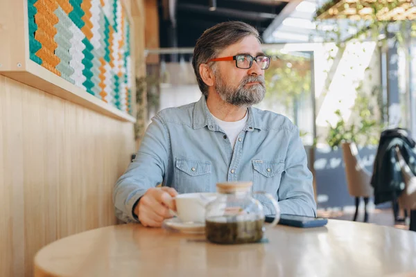 Bearded senior man savoring a warm cup of tea in a modern cafe, gazing thoughtfully out the window while enjoying a peaceful moment