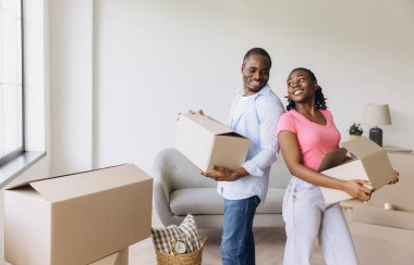 Smiling African American couple joyfully carrying cardboard boxes while moving into their new apartment, embracing fresh beginnings together