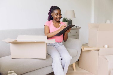 African american woman sitting on a couch, organizing and writing on a clipboard amidst moving boxes in a bright, modern living room