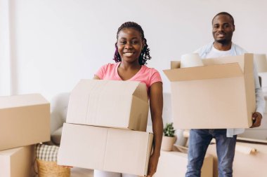 Young african american couple carrying cardboard boxes moving in new house, smiling and looking at camera