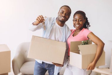Smiling African American couple moving into a new apartment, carrying cardboard boxes and keys, excited for their fresh start together