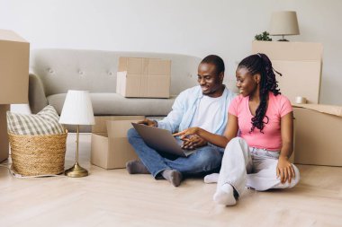 Young african american couple sitting on floor among cardboard boxes using laptop and planning new home interior design while moving into new apartment