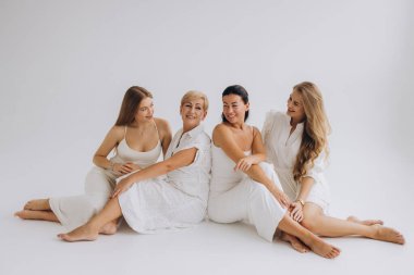 Four cheerful women of different ages wearing white dresses are sitting together on the floor against a white background, creating a serene and elegant atmosphere