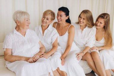 Five women of different ages, possibly representing a family, are sitting together on a sofa, dressed in white, engaging in conversation and displaying positive emotions