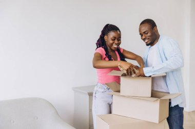 Young smiling african american couple unpacking belongings from cardboard boxes in new apartment after moving in