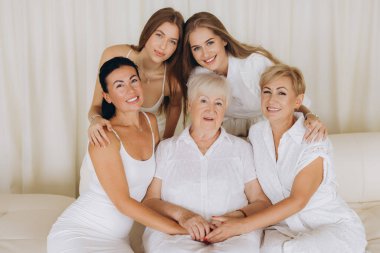 Five women of different generations, wearing white dresses, are smiling and holding hands, representing family unity and connection
