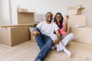 Young african american couple sitting on the floor of their new apartment, among cardboard boxes, showing keys and smiling