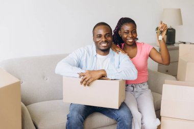 Excited couple showing keys to their new apartment while sitting on sofa surrounded by cardboard boxes, happy to start new life chapter