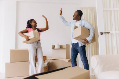 Young black couple giving high five while carrying cardboard boxes, celebrating moving into new home