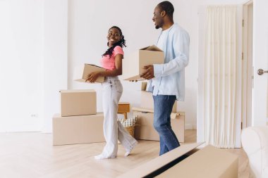 Young, smiling African American couple joyfully carrying cardboard boxes while moving into their new home, embracing a fresh start together