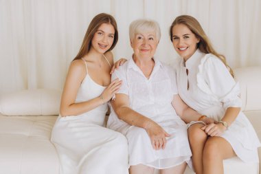 Three generations of women, dressed in elegant white dresses, sitting together on a plush white sofa, sharing joyful smiles and warmth