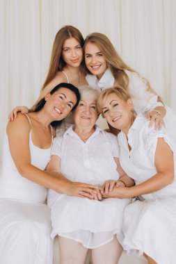 Five women of different ages wearing white dresses are showing affection and love to their grandmother