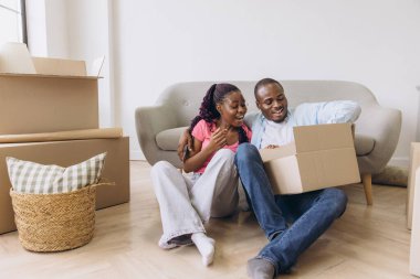 Young african american couple is unpacking cardboard boxes in new apartment, smiling and embracing