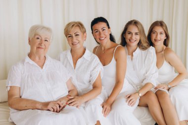 Five women representing different generations of a family are sitting together on a white sofa, wearing white clothes, creating a sense of unity and connection