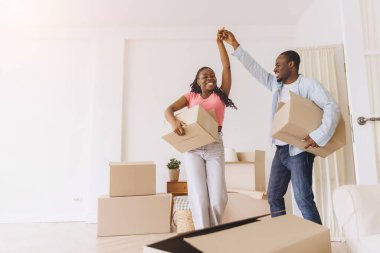 Happy african american couple dancing and holding cardboard boxes, celebrating moving to new home