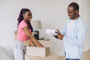 Happy couple unpacking belongings from cardboard boxes, man writing on clipboard, moving in new house