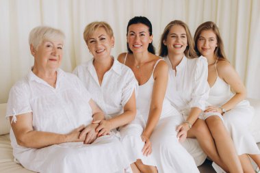 Five women of different ages, grandmother, mother and daughters, wearing white dresses, are sitting on a white sofa, smiling and looking happy