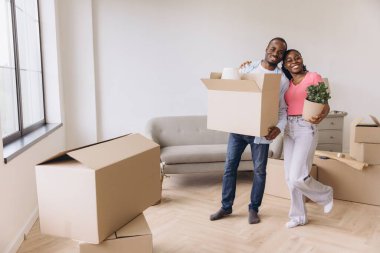Young black homeowners joyfully carrying boxes and a potted plant into a bright, spacious new home, smiling and embracing each other