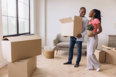 Young black couple moving into a new home, carrying boxes and a houseplant, smiling and happy about their new life together