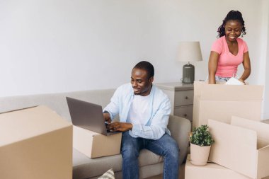 Happy African American couple moving into a new apartment, unpacking boxes and using a laptop to organize their belongings together