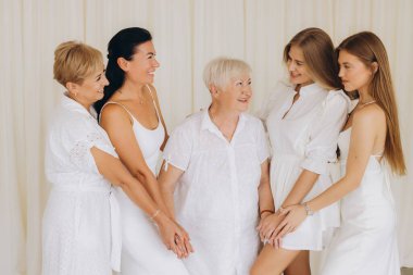 Four generations of women holding hands, enjoying each other's company, smiling joyfully while dressed elegantly in white attire