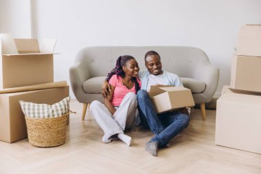 Young african american couple sitting on the floor and unpacking cardboard boxes in new apartment