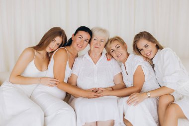 Five women of different generations wearing white dresses sitting together on a white sofa, showing love, connection and family unity