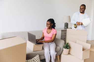 Happy african american couple moving into new apartment, woman using laptop and man unpacking boxes