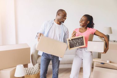 Smiling African American couple carrying cardboard boxes and a moving day sign while entering their new home, embracing the excitement of fresh beginnings