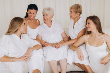 Four women of different generations, dressed in white, sit together on a sofa, creating a heartwarming scene of family bonding