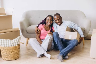 Young black couple sitting on the floor in their new apartment surrounded by cardboard boxes, hugging and smiling