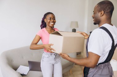 Smiling african american woman receiving cardboard box from male mover, delivering belongings to new home