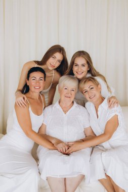 Five women of different ages, dressed in white, embracing and smiling, representing family bonds and unity