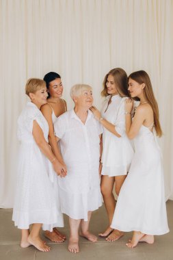 Four women of different generations wearing white dresses are standing together, smiling and embracing, symbolizing family unity and love