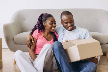 Smiling African American couple joyfully unpacking belongings from cardboard boxes in their new home, embracing a fresh start together