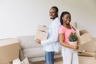 Young couple smiling while carrying cardboard boxes and a houseplant into their new apartment, embracing a fresh start together